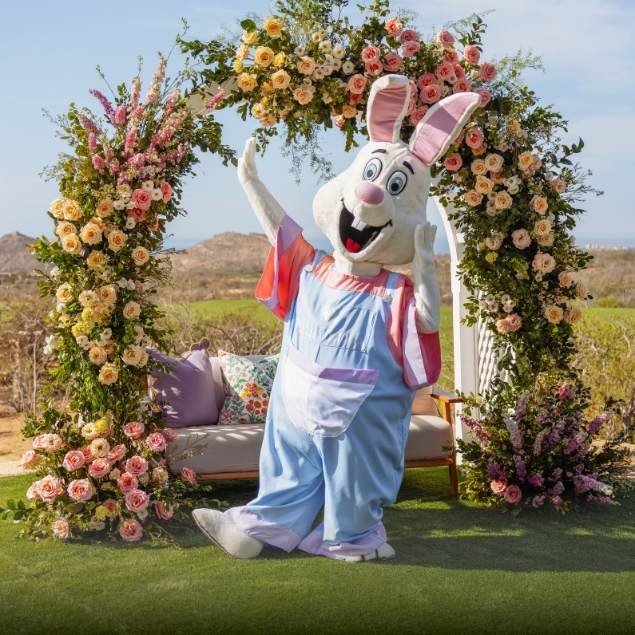 Easter Bunny mascot posing beneath a floral arch with spring blooms and outdoor seating at a festive Easter celebration event in Los Cabos.
